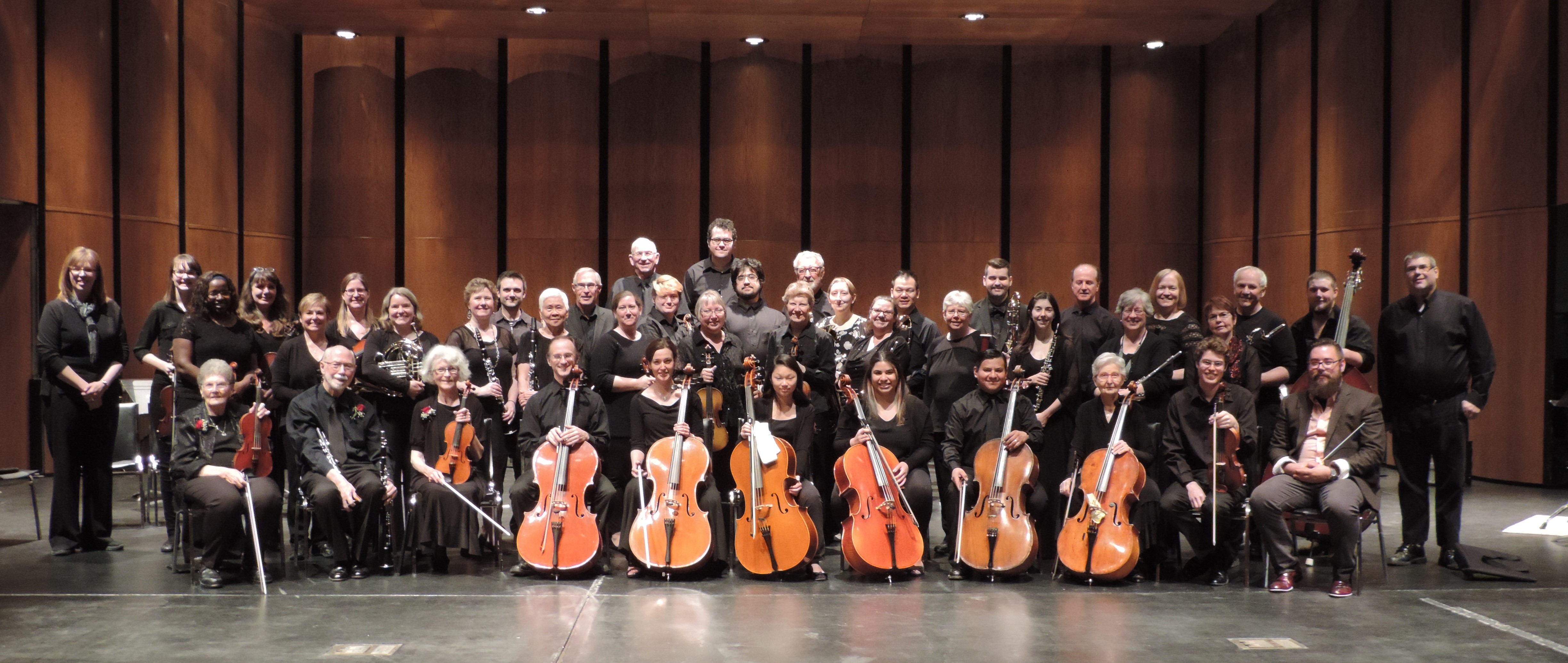 Musicians sit and stand in rows hoolding instruments, on a large stage.