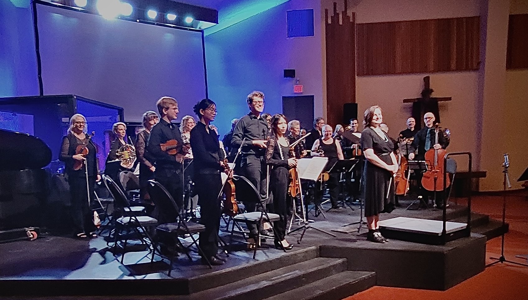 Musicians stand holding instruments on a raised stage under bright lights. A conductor in a black dress smiles to acknowledge applause. 